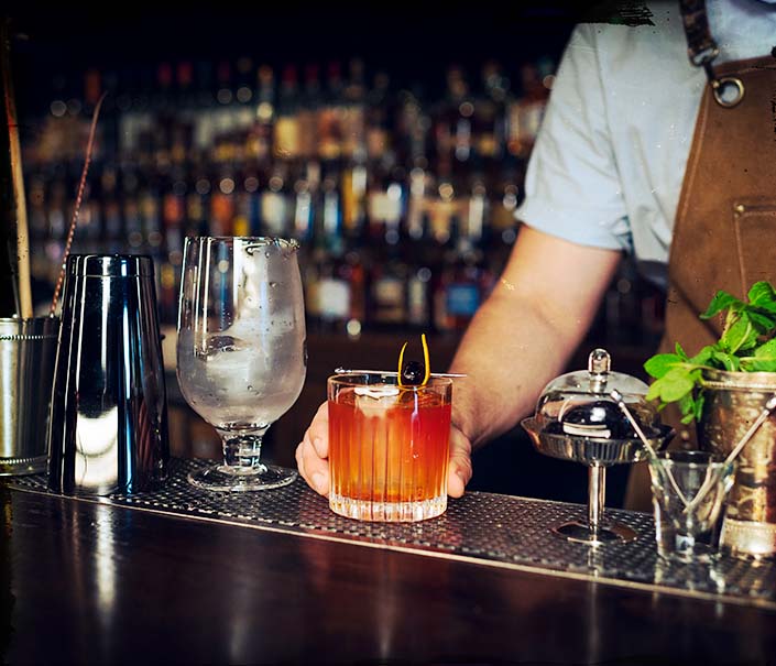 Bartender serving red brown Labyrinth whiskey cocktail with orange peel and cherry garnish at bar - Redemption Whiskey